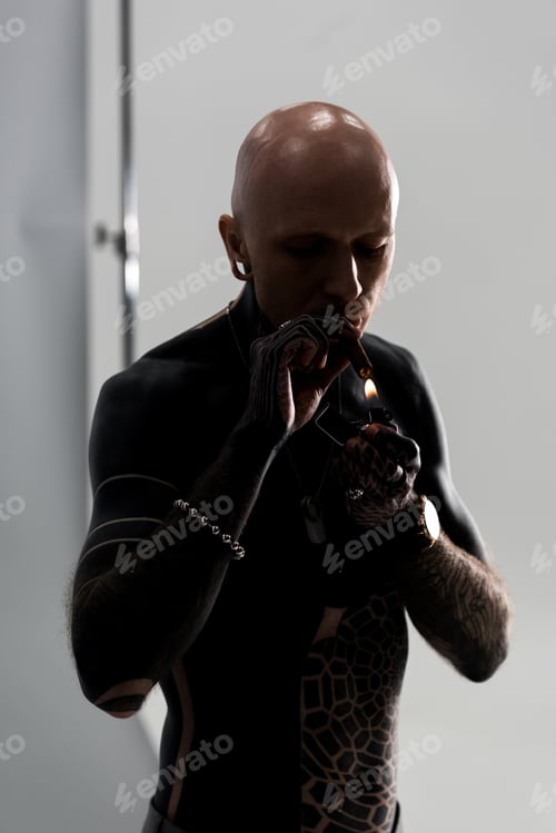 Preview: bare-chested bald man with tattoos holding lighter and smoking cigar on grey