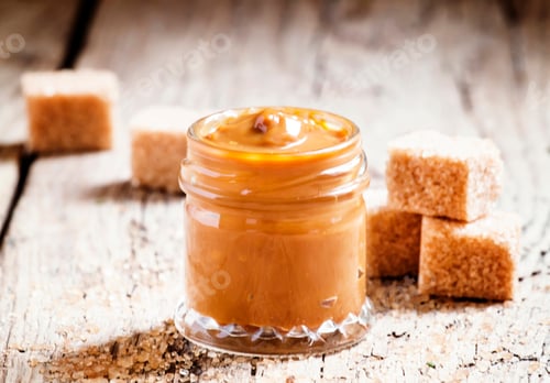 Preview: Sweet liquid caramel in a small jar and brown cane sugar, on the old wooden background