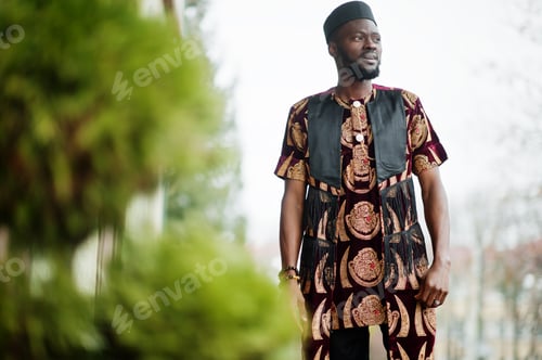 Preview: African stylish and handsome man in traditional outfit and black cap standing outdoor.
