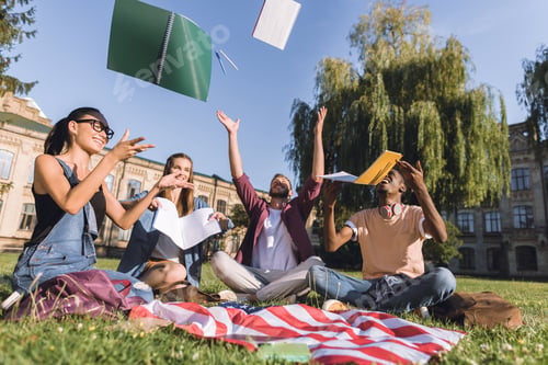 Preview: multiethnic cheerful students throwing notebooks while sitting on grass