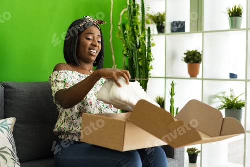 Preview: African young woman sit on couch at home unpack cardboard box with online purchase