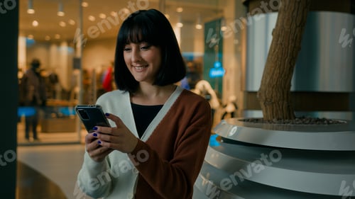 Preview: Smiling 20s woman lady female girl indoors in shopping mall sitting looking at mobile phone screen