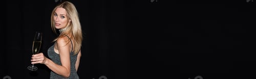 Preview: Trendy woman in dress holding champagne and looking at camera isolated on black, banner