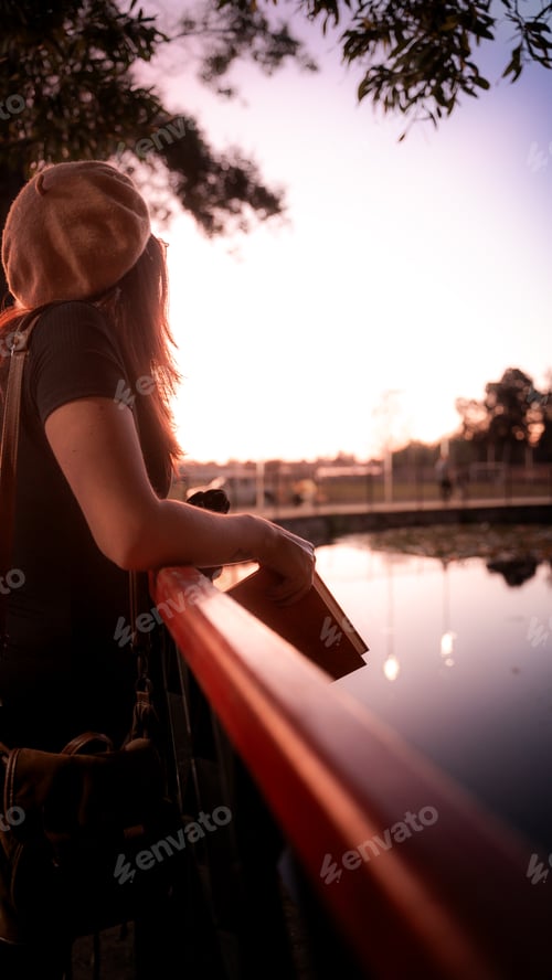 Preview: Young girl reading a book on a bridge at sunset in a park with trees