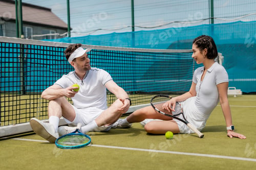 Preview: happy man and woman in sportswear sitting on tennis court, rackets and balls, summer, couple sport