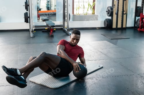 Preview: African American man performing twists wearing red shirt shorts on mat at gym holding medicine ball