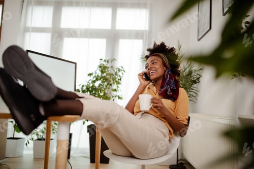 Preview: Black designer relaxing on chair in office and chatting on smartphone with legs on the table.