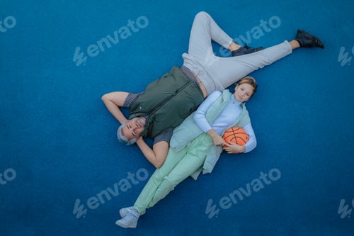 Preview: Top view of happy father and teen daughter lying and looking at camera outside at basketball court.