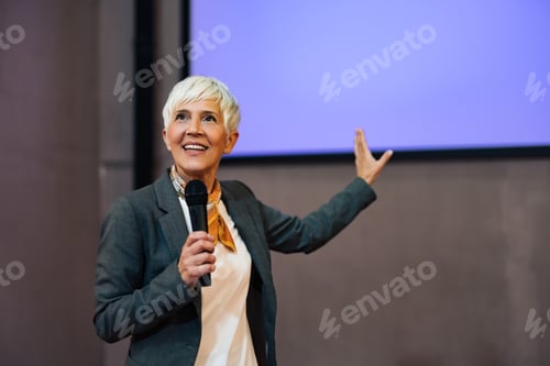 Preview: A smiling woman giving a speech, pointing at the projection screen, elegantly dressed.