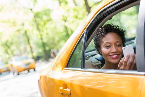 Preview: A woman sitting in the rear passenger seat of a yellow cab, checking her smart phone.