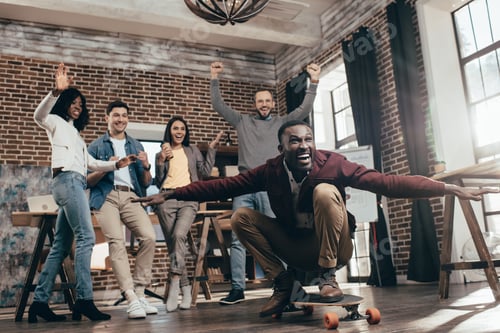 Preview: group of happy multiethnic coworkers having fun with skateboard in loft office