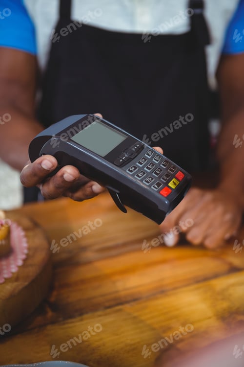 Preview: African American woman holding out card terminal at wooden cafe counter, with pink cake stand