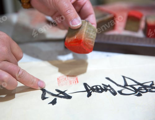 Preview: Hand holding wooden stamp with red paint, marking white piece of paper