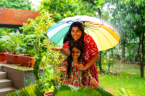 Preview: Two Indian girls playfully enjoy monsoon rain under umbrella, sharing joyful outdoor moment