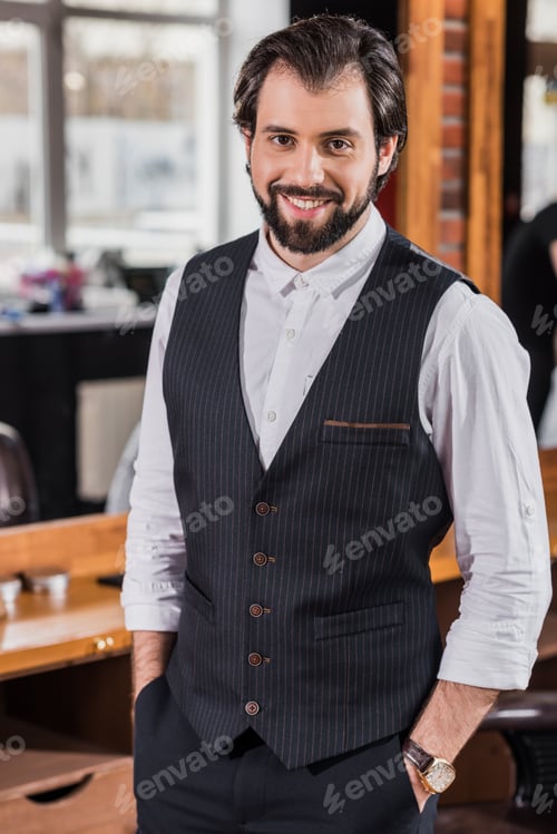 Preview: handsome young barber in stylish vest looking at camera in barbershop