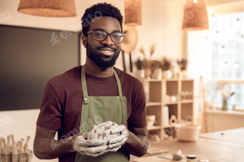 Preview: Dark-skinned smiling sculptor with wet clay in hands looking contented
