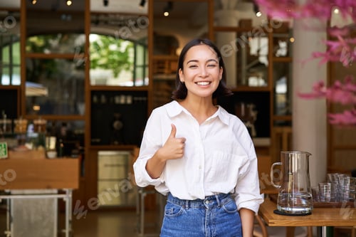 Preview: Confident asian businesswoman, showing thumbs up, standing near entrance of her cafe or restaurant