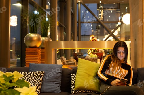 Preview: Young woman in cosy sweater sitting in hotel lounge using mobile phone