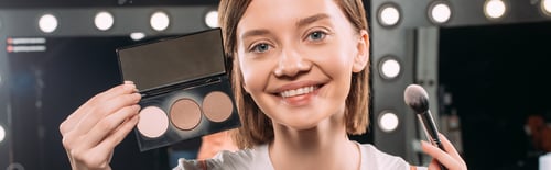 Preview: Panoramic crop of girl smiling at camera while holding makeup set and cosmetic brush in photo studio