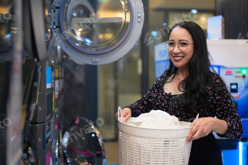 Preview: Woman smiling while holding laundry basket near industrial washing machines in public laundromat