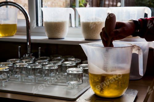 Preview: Close up of person holding jug, stirring liquid wax for candle making with wooden spoon.