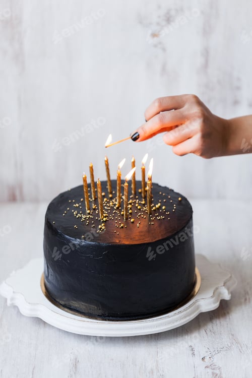 Preview: Black birthday cake with woman hand burning candles, white background
