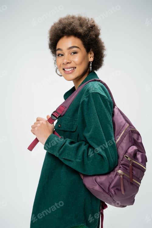 Preview: cheerful african american woman in shirt and hoop earrings holding backpack and looking at camera