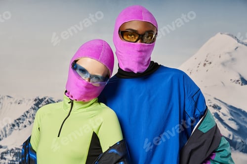 Preview: young stylish couple in warm vibrant outfits and pink balaclavas with snowy backdrop, winter concept