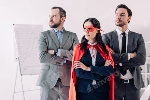 Preview: super businesswoman standing with crossed arms in mask and cape with businessmen in office