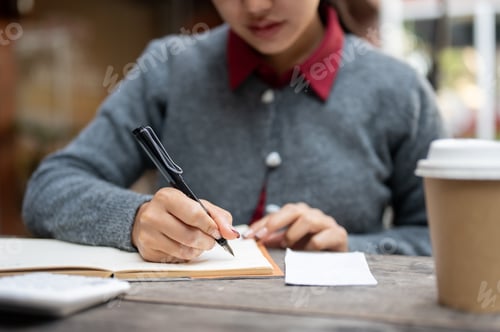 Preview: A cropped image of a woman writing or taking notes in a book while sitting at a table outdoors.