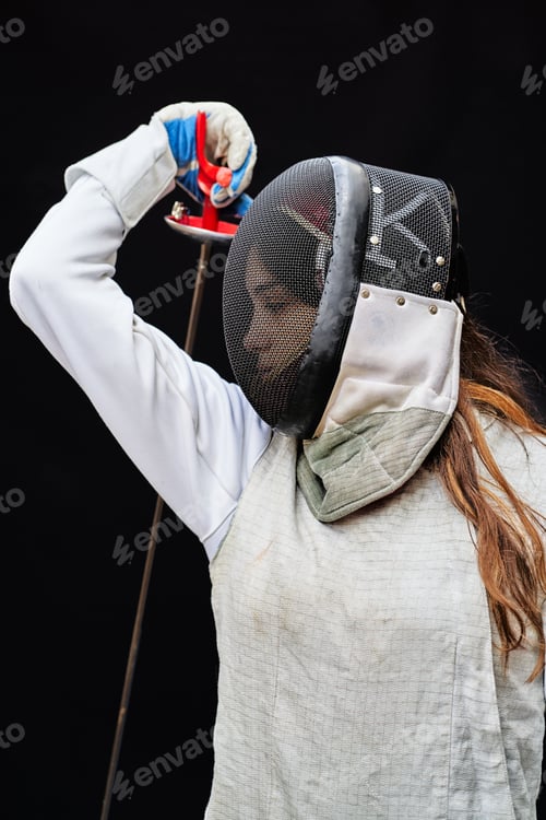 Preview: Portrait of woman dressed in white fencing costume practicing with the sword on black background.
