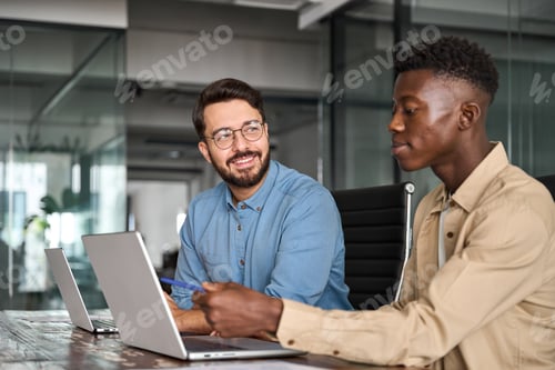 Preview: Two busy happy diverse male employees using laptops working talking in office.