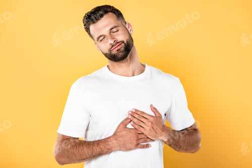 Preview: pleased handsome man in white t-shirt touching heart isolated on yellow
