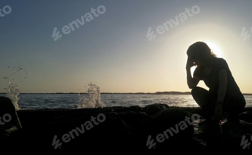 Preview: Silhouetted babyboomer sitting on rocks at seashore thinking about psychology and her future.