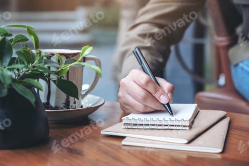 Preview: Closeup image of a woman's hand writing on blank notebook with coffee cup on wooden table