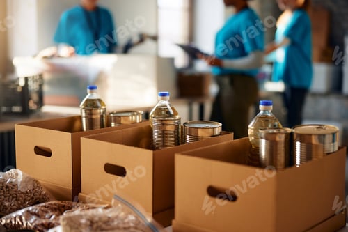 Preview: Donation boxes with sorted groceries at food bank with volunteers in the background.