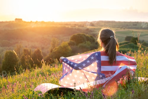 Preview: Young woman with USA national flag standing outdoors at sunset.