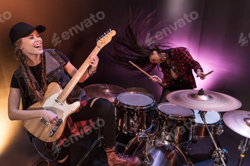Preview: Excited young women with drums set and electric guitar performing rock concert on stage