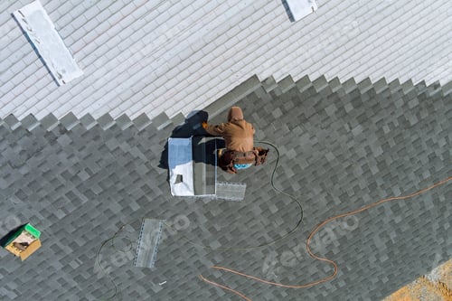 Preview: A worker uses an air hammer to nail new roof shingles to bitumen using a top view from above