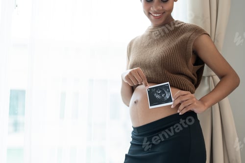 Preview: Happy African American pregnant woman showing ultrasound scan picture of her unborn at home