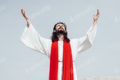 Preview: Jesus in robe, red sash and crown of thorns standing with raised hands and praying in desert