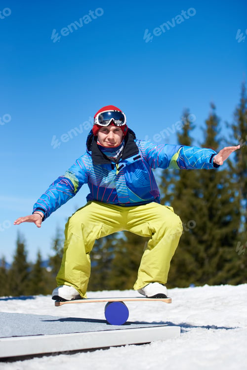 Preview: Male snowboarder balancing on snowboard in white snow on background of blue sky and spruce trees.