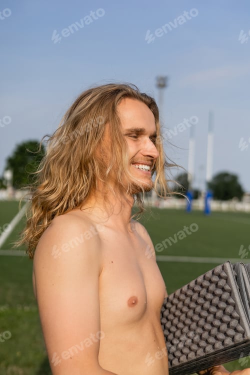 Preview: overjoyed shirtless man with long hair holding yoga mat and smiling outdoors
