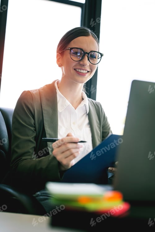 Preview: Young businesswoman with glasses working on document and laptop computer. Smiling to camera.