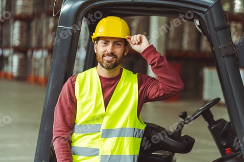 Preview: Bearded man, driver, wearing helmet, standing near forklift, looking at camera standing in warehouse
