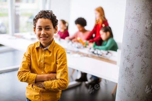 Preview: Cute little boy standing in front of kids programming electric toys and robots at robotics classroom