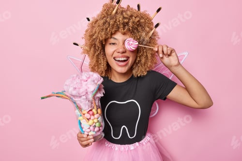 Preview: Positive tooth fairy poses with sweet colorful candies covers eye with lollipop on stick dressed in