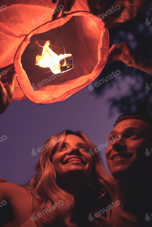 Preview: low angle view of smiling young couple launching sky lantern on river beach in evening