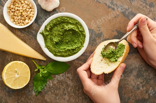Preview: cropped view of woman adding pesto sauce on baguette slice near fresh ingredients on stone surface