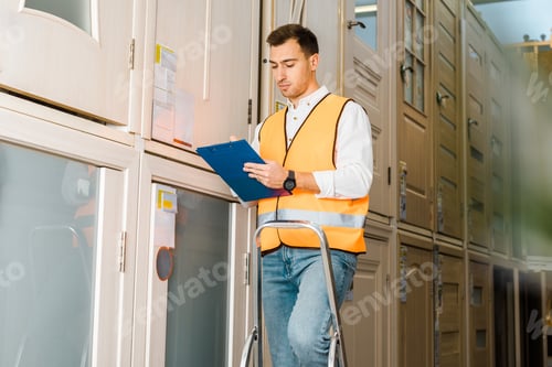 Preview: selective focus of concentrated worker with clipboard standing on ladder in warehouse